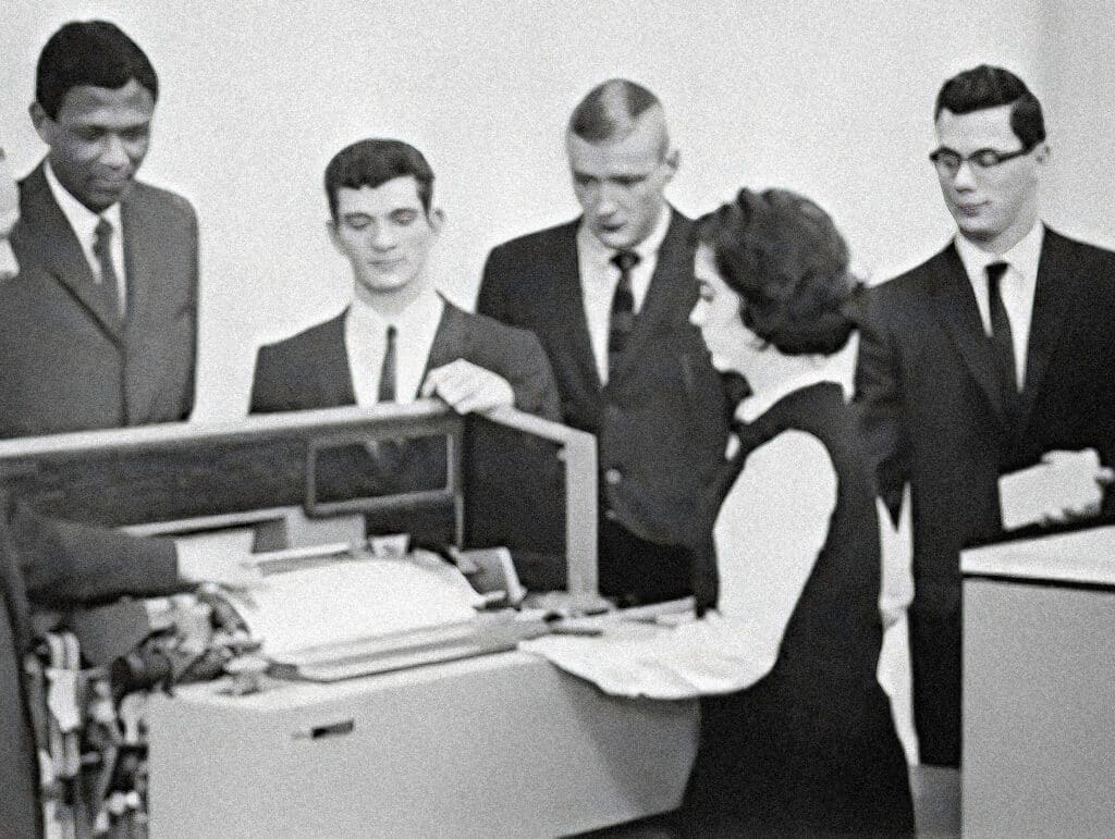A group of men in suits watch a woman operate a large office machine in a black-and-white photo.