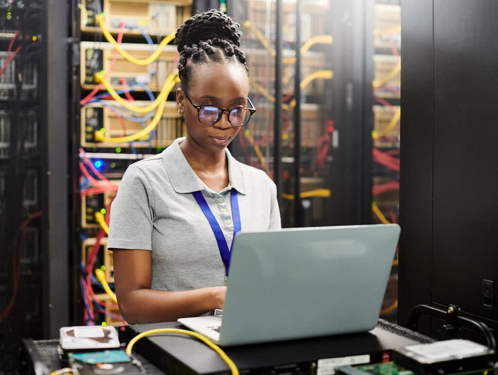 A woman in a server room works on a laptop, surrounded by racks of servers and colorful network cables.