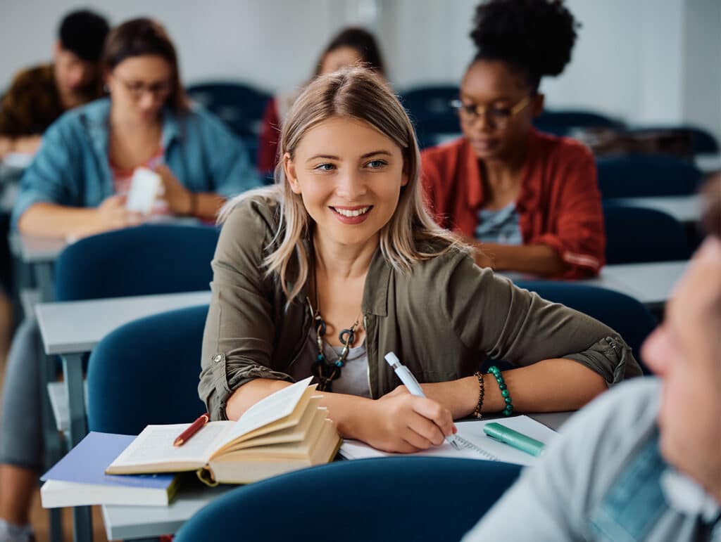 A smiling woman with blonde hair takes notes during a class, sitting among other focused students in a classroom.