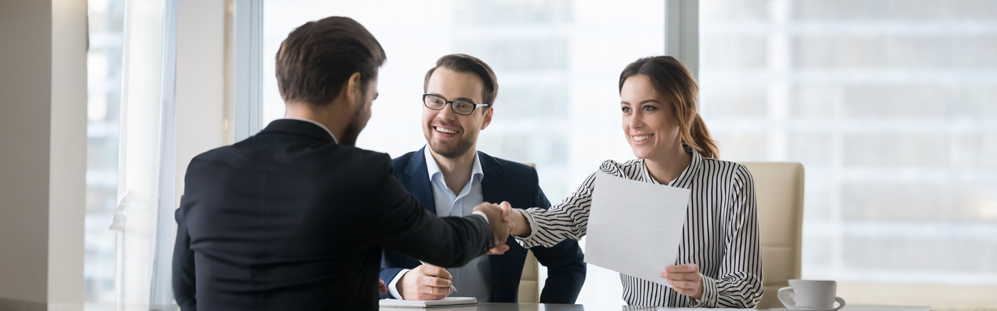 Person in a business suit shaking hands with two smiling interviewers across a desk in an office setting.