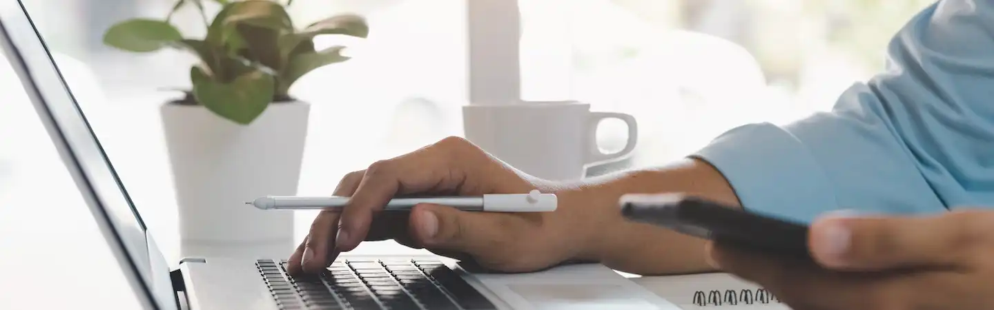 Person holding a pen and a smartphone while working on a laptop, with a coffee cup and plant in the background.