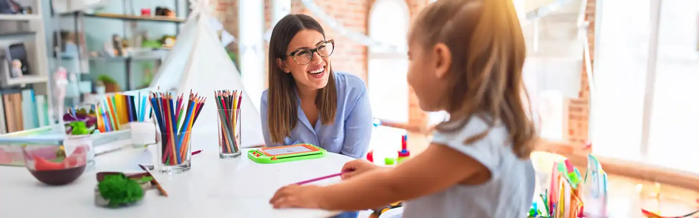 A woman with glasses smiles and interacts with a young girl in a well-lit art classroom filled with colorful supplies.