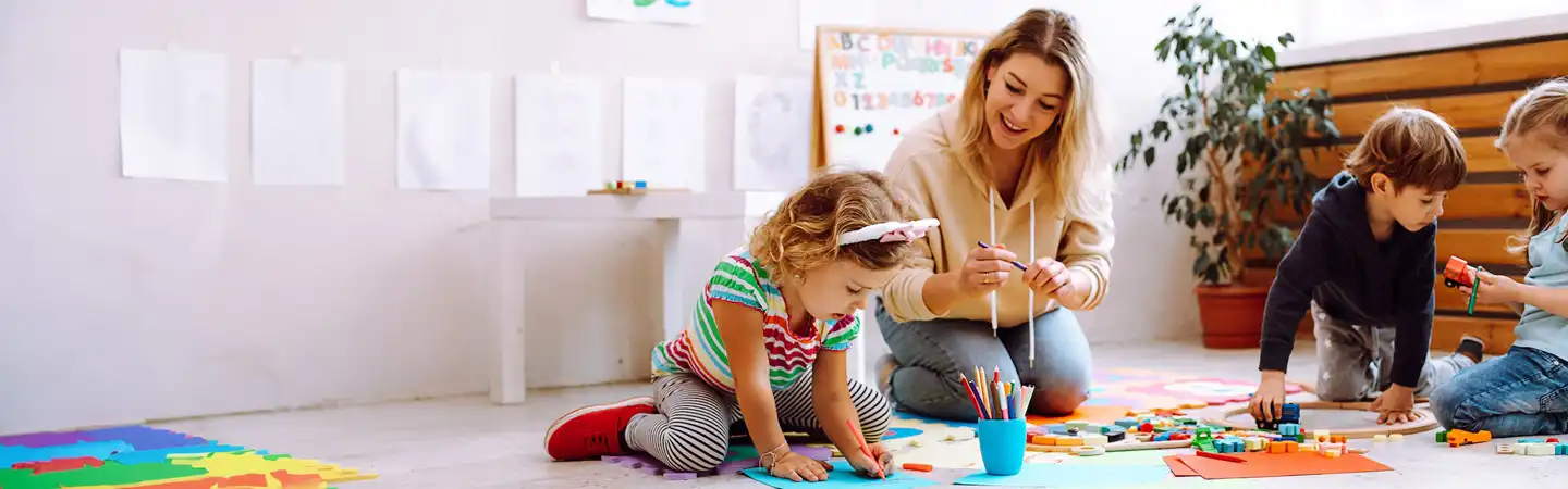 Adults and children engaged in various creative activities in a bright, colorful classroom with art supplies scattered around.