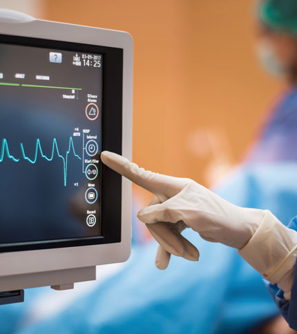 A gloved hand touches the controls on a medical monitor displaying a heartbeat and other vital signs. Blurred background shows medical personnel in scrubs, suggesting a hospital or surgical setting.
