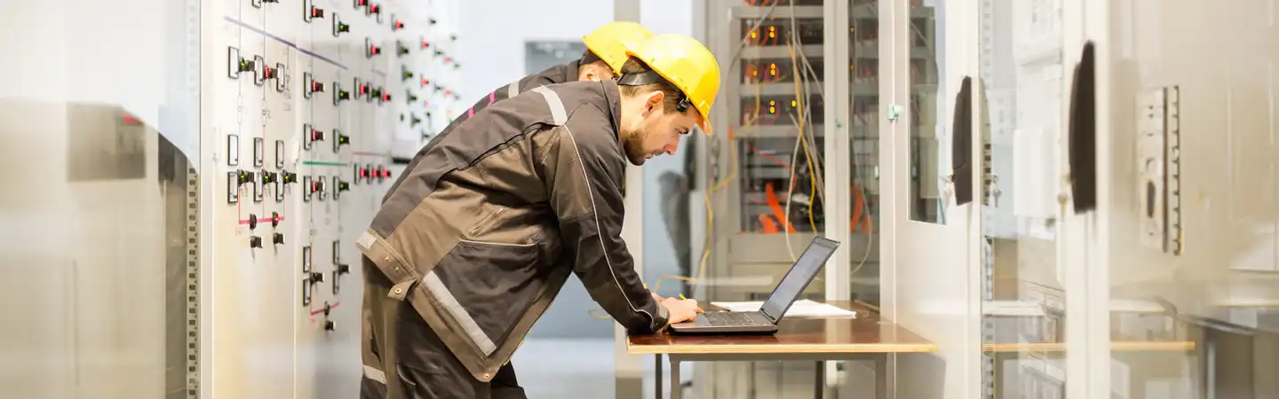 Two workers in safety gear operate a laptop in a control room with panels and electronic equipment.