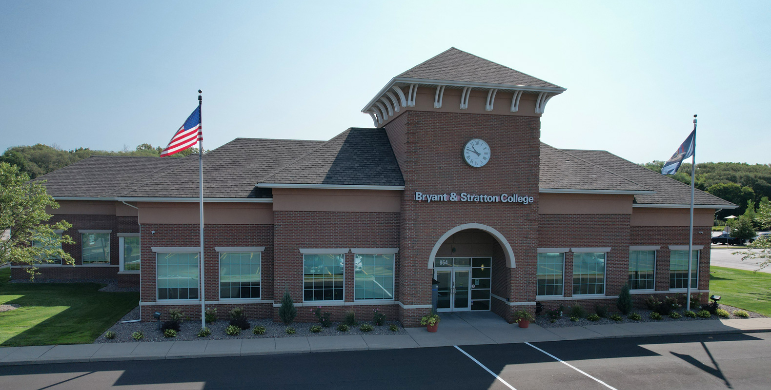 A brick building with a clock tower and two flagpoles in front, displaying the signs "Bryant & Stratton College." The entrance has glass doors, and manicured landscaping lines the sidewalk.