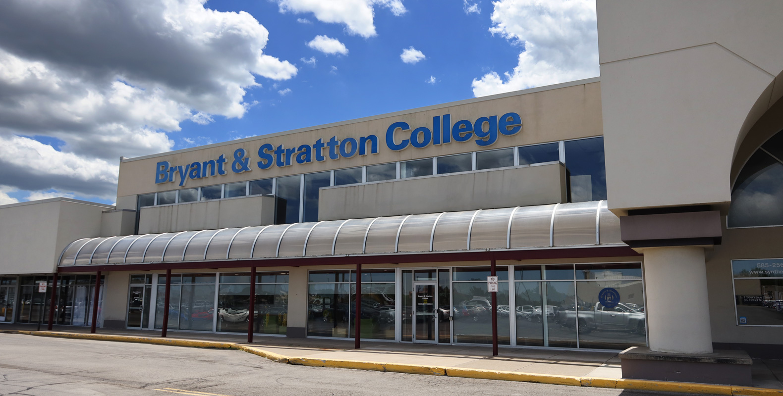 The exterior of the Bryant & Stratton College building in Henrietta, NY features large blue letters above glass doors and windows, set against a partly cloudy sky in the background.