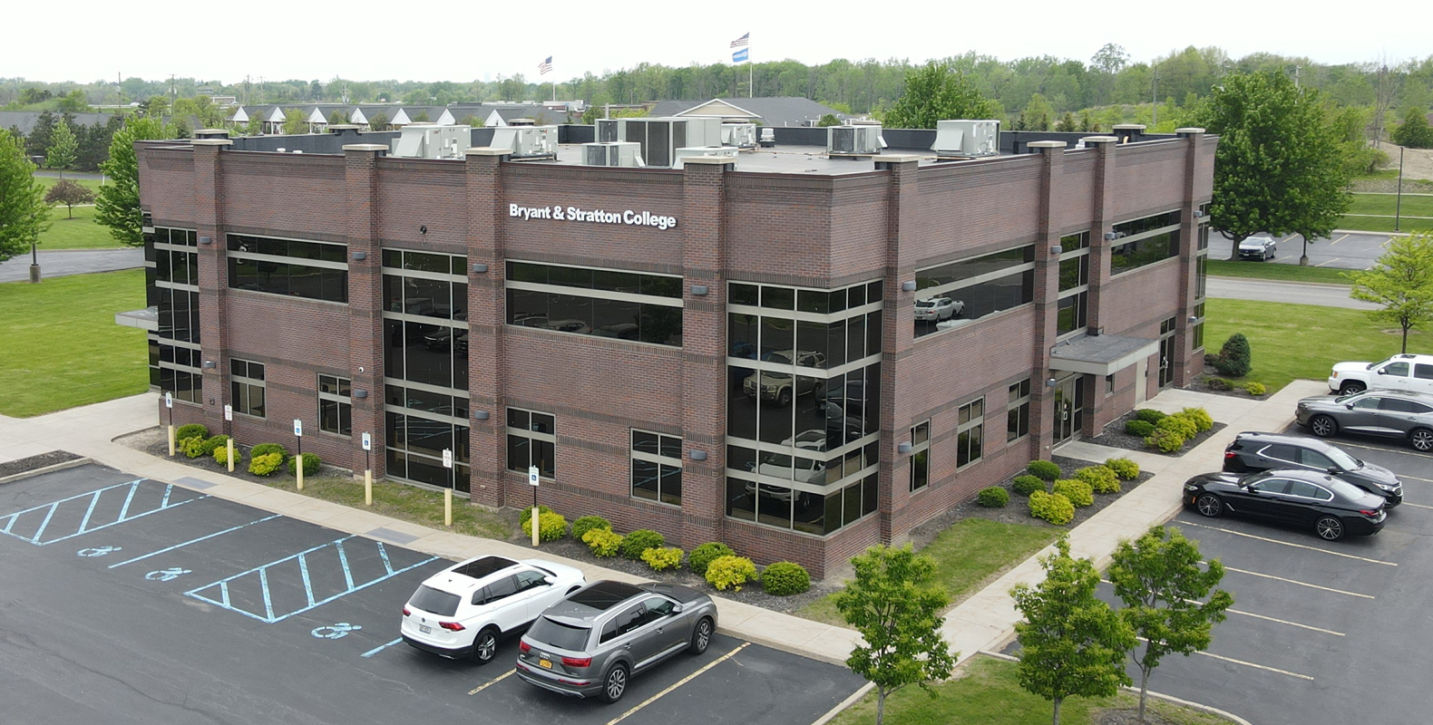 A two-story brick building with large windows shows a sign reading "Bryant & Stratton College" in Orchard Park, NY. Several cars are parked outside, including accessible spaces. Green trees and grass surround the building.