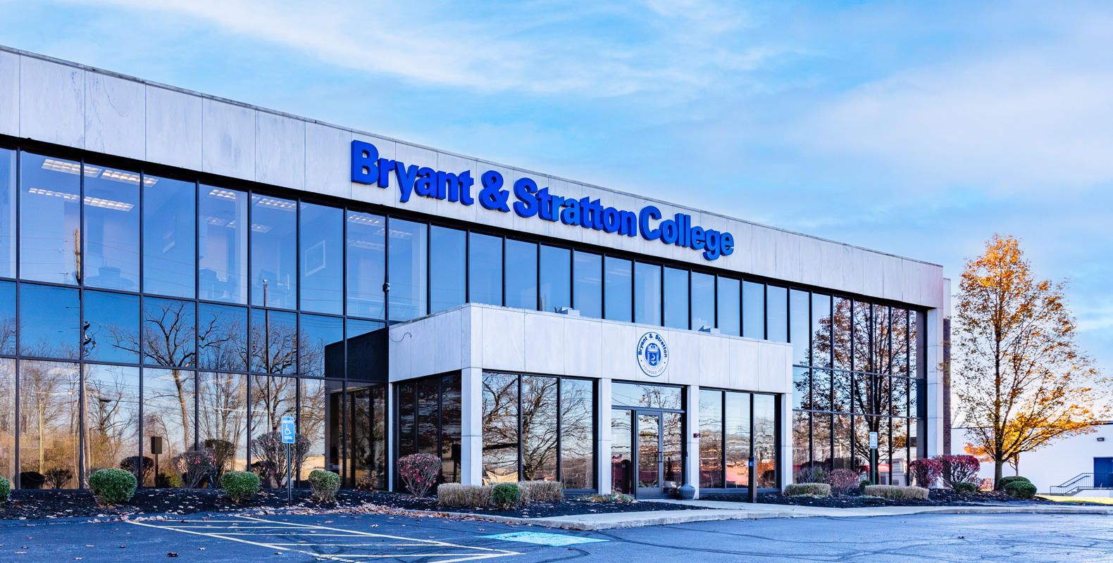 The image shows the exterior of Bryant & Stratton College, a modern two-story building with large reflective windows, a blue sign, and a parking lot in front. Trees and bushes surround the entrance.