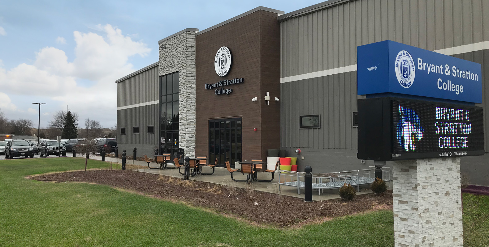 Exterior view of Bryant & Stratton College building with logo. There are benches and tables near the entrance, a digital sign displaying the school name, and cars parked in the lot on a cloudy day.