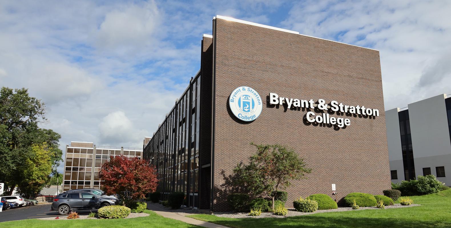 A large brick building with the Bryant & Stratton College logo and name on its exterior, representing one of the prominent Syracuse colleges, surrounded by green shrubs, trees, a sidewalk, and parked cars under a partly cloudy sky.