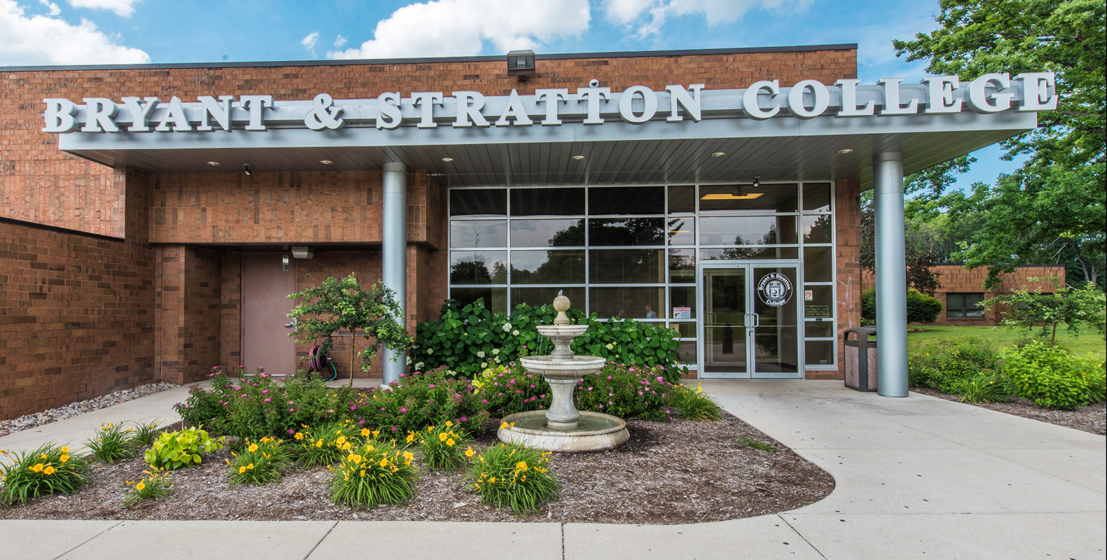 The entrance of Bryant & Stratton College in Wauwatosa, WI features large glass windows, brick walls, a double-door entrance, a decorative fountain, and vibrant flower beds. The college name is prominently displayed above the entrance.