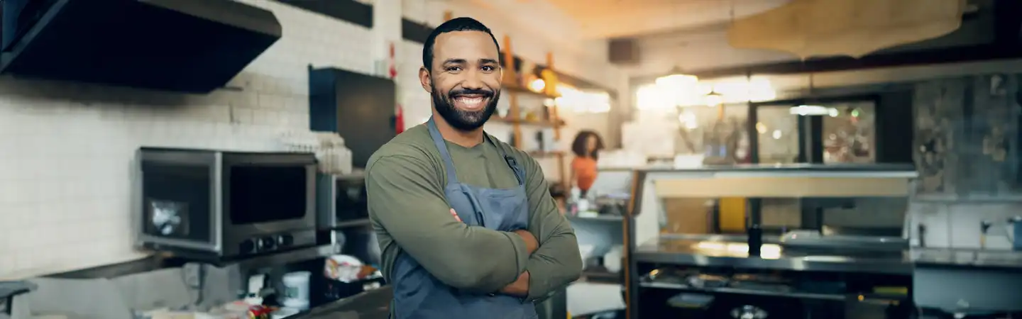 A smiling man in an apron stands with arms crossed in a cozy, well-lit kitchen.