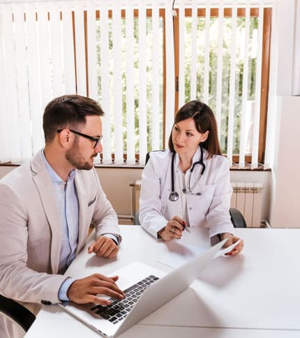 A doctor and a patient are seated at a desk in a bright office. The doctor, wearing a white coat and stethoscope, is attentively listening while holding a pen. The patient is using a laptop and appears engaged in conversation.