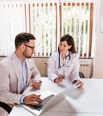 A doctor in a white coat and stethoscope talks to a man in a suit at a desk. The man uses a laptop while they both review a document. They are in a bright office with large windows and vertical blinds.