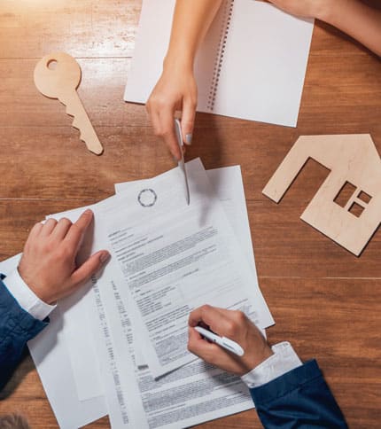 People signing real estate documents on a wooden table with a large key and house-shaped cutout nearby.