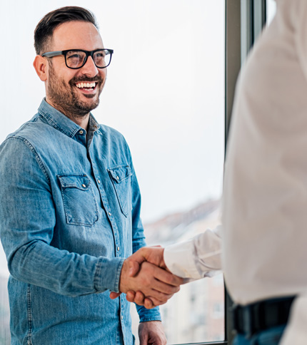 Smiling man in glasses and a denim shirt shaking hands with another person indoors near a bright window, exemplifying the welcoming spirit of a Human Resources Assistant.