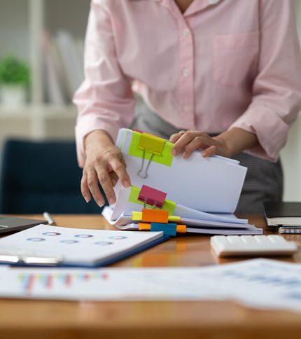 An Information Clerk in a pink shirt organizes stacks of documents with colorful binder clips on a desk, which also has charts, a keyboard, and office supplies.