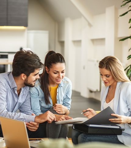 Three people sit together in a bright office, looking at documents and smiling. An Insurance Reimbursement Specialist holds a folder and pen, discussing important details with a man and another woman seated beside her.