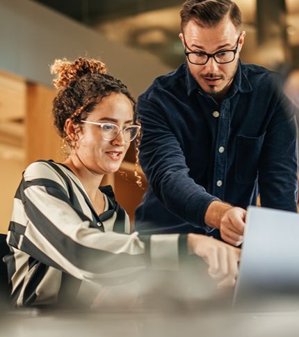 Two colleagues, including an IT Coordinator, work together at a desk, looking intently at a laptop screen. One is seated and typing while the other stands beside them, pointing at the screen and offering guidance. Both wear glasses and appear focused.