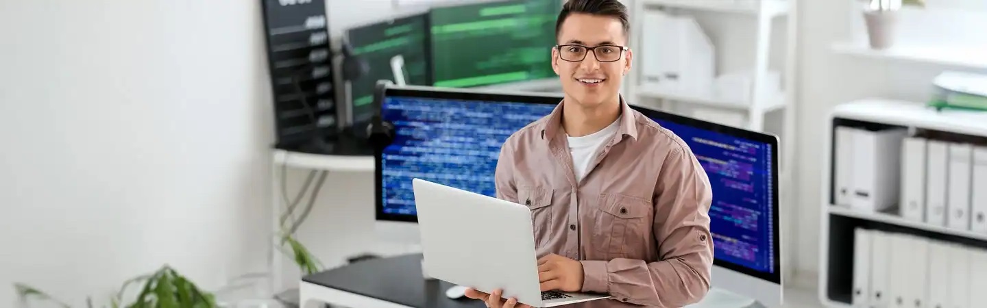 A person in glasses sits at a desk with a laptop, several monitors displaying code in the background, smiling at the camera.