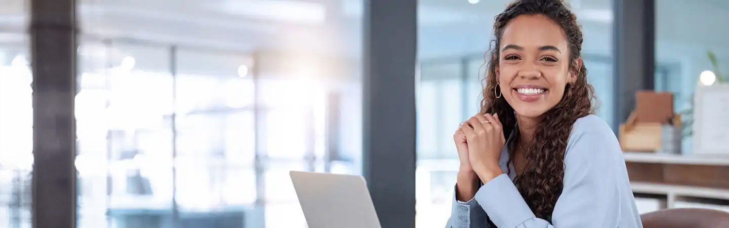 Smiling person with curly hair sits at a desk with a laptop in a bright office environment.
