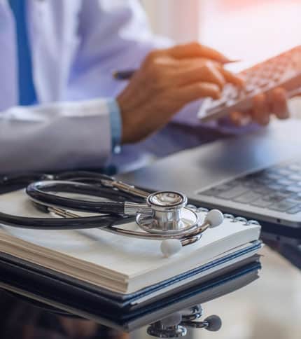 A doctor sits at a desk, using a calculator and a laptop. A stethoscope and a notepad are placed on the desk, suggesting a medical setting.
