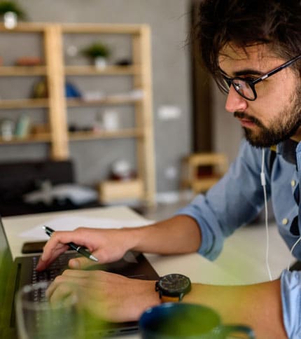 A man with glasses and earphones is focused on typing on a laptop at a desk. A notebook and pen are nearby. The background features a wooden shelf with books and plants. The setting appears to be an office or study area.