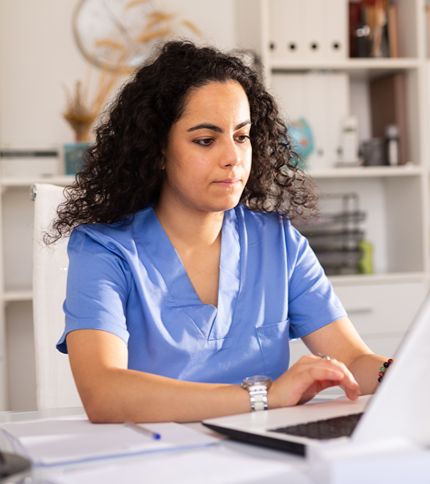 A Medical Office Assistant with curly hair, dressed in blue scrubs, sits at a desk working on a laptop in a well-lit office environment.