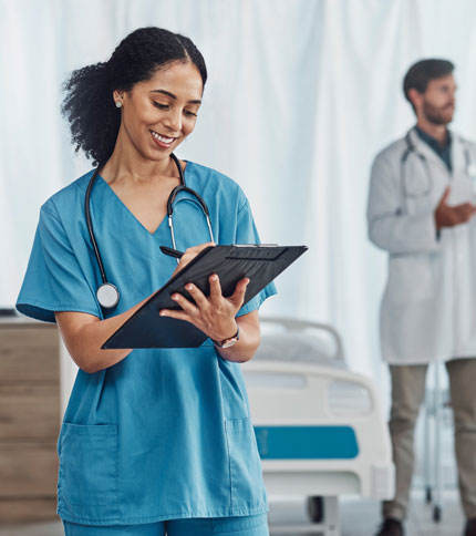 A healthcare professional in blue scrubs writes on a clipboard, smiling. She has a stethoscope around her neck. In the background, a blurred figure in a white coat stands near a hospital bed.