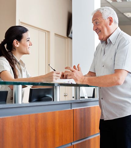 An older man smiles and talks with a young healthcare receptionist at a modern front desk. The receptionist is seated, holding a pen, and listens attentively, both appearing friendly and engaged in the medical office setting.