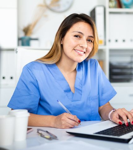 A health information technician in blue scrubs sits at a desk, smiling while holding a pen and working on a laptop. Papers, a coffee cup, and office supplies are on the desk, with shelves and office items in the background.