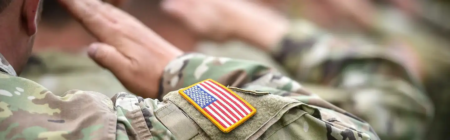 Soldiers in camouflage uniforms saluting, with a focus on a U.S. flag patch on one soldier's sleeve.