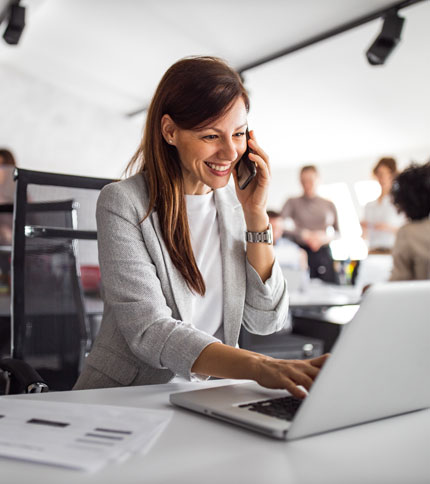 A smiling woman in a gray blazer talks on a smartphone while working on a laptop in a bright office. Papers are on the desk, and other people are blurred in the background.