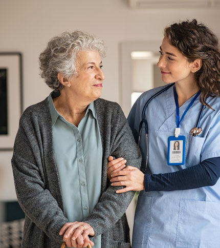 A nurse in blue scrubs and a name tag supports an elderly woman with gray hair and a walking cane. They are smiling at each other in a warmly lit room, conveying a sense of care and trust.