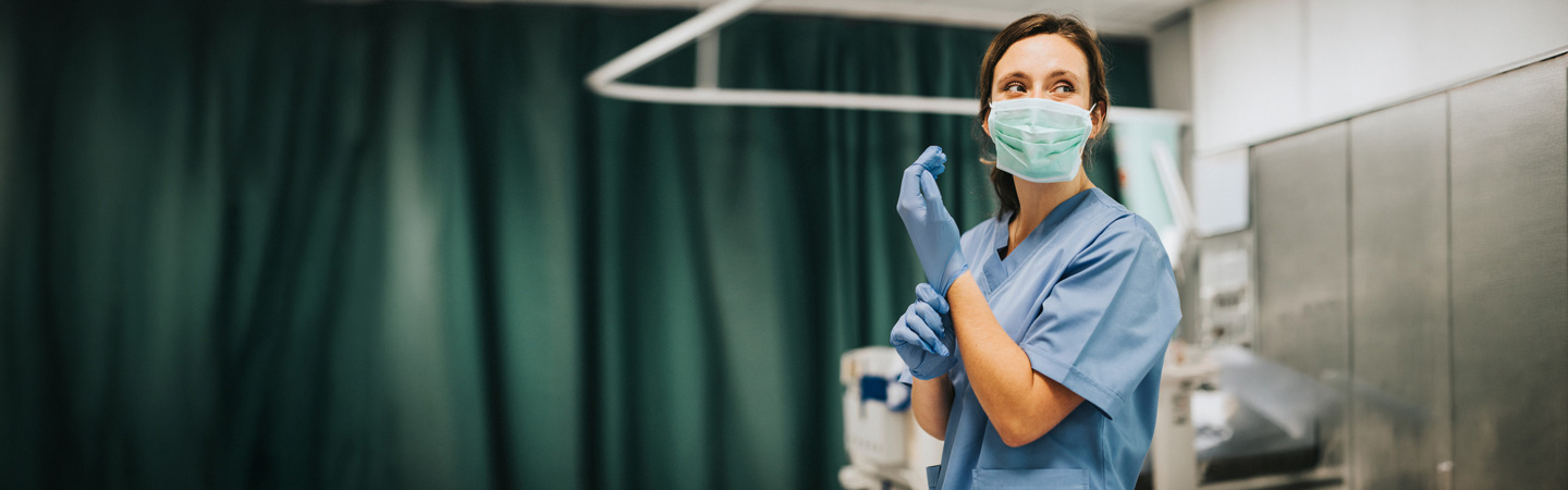 A healthcare worker wearing scrubs, a surgical mask, and gloves stands in a hospital room with green privacy curtains, preparing for a medical procedure.