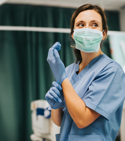 A healthcare worker wearing blue scrubs, a medical mask, and blue gloves stands in a clinical setting, adjusting one glove while looking to the side.