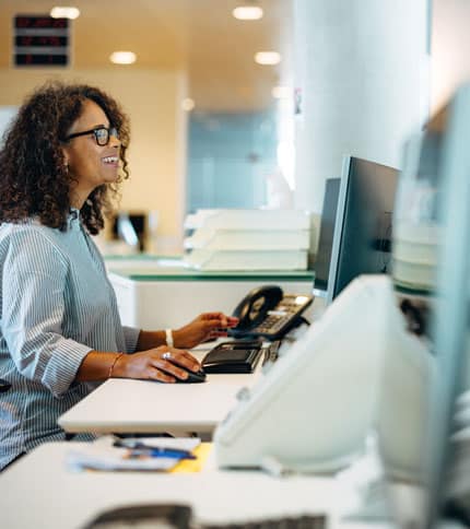 A person with curly hair and glasses is sitting at a desk and using a computer. They are smiling and have a phone nearby. The setting appears to be an office environment with stacks of paper in the background.