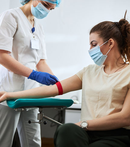 A healthcare professional wearing a mask and gloves prepares to draw blood from a seated woman with a red tourniquet on her arm. Both are wearing masks in a clinical setting.