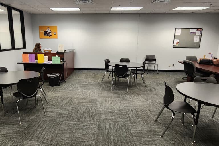 A classroom with round tables and chairs, a small reception desk with a staff member, and bulletin boards on the walls.