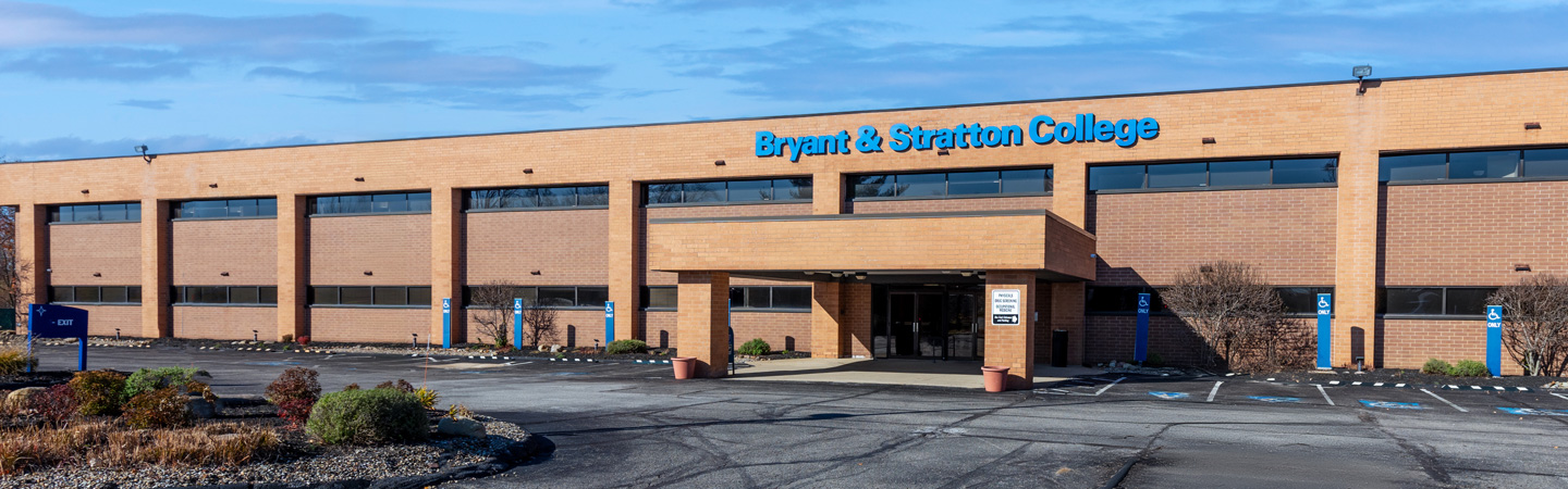 A brick building with the sign "Bryant & Stratton College" above the main entrance surrounded by parking spaces and plants.