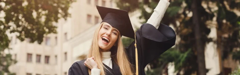 A graduate in a cap and gown celebrates outdoors, holding her diploma and raising her arm joyfully.