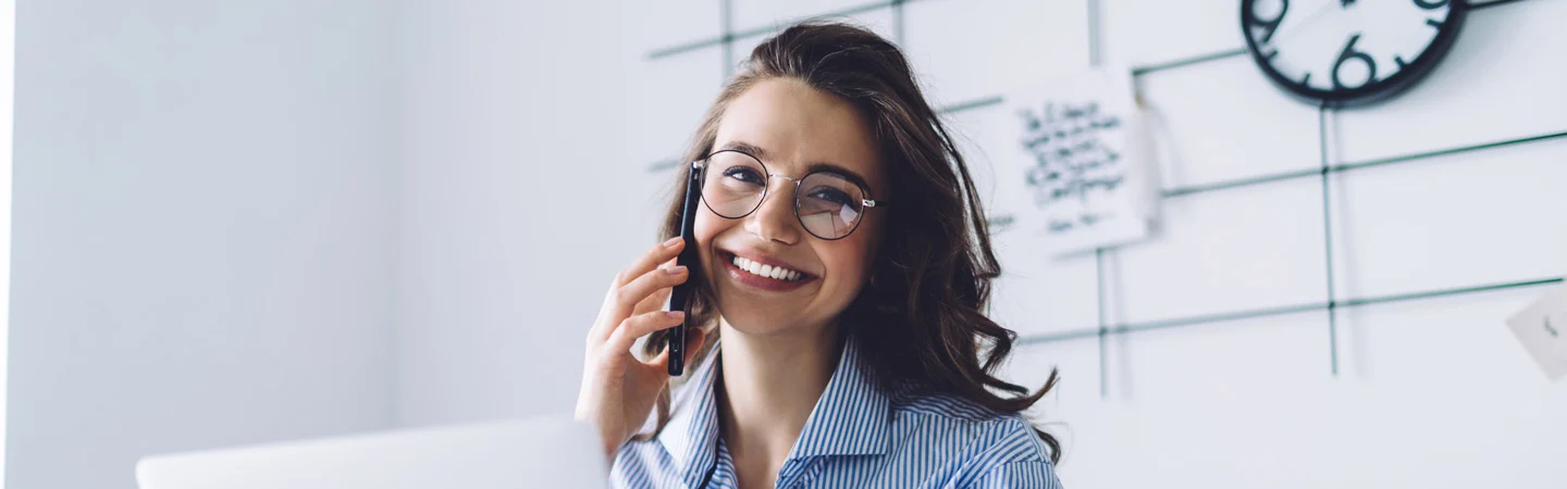 Smiling person with glasses talks on the phone in a bright office with notes and a clock on the wall.