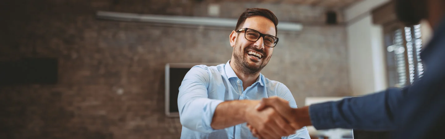 A man in glasses smiling while shaking hands with another person in a modern office setting.