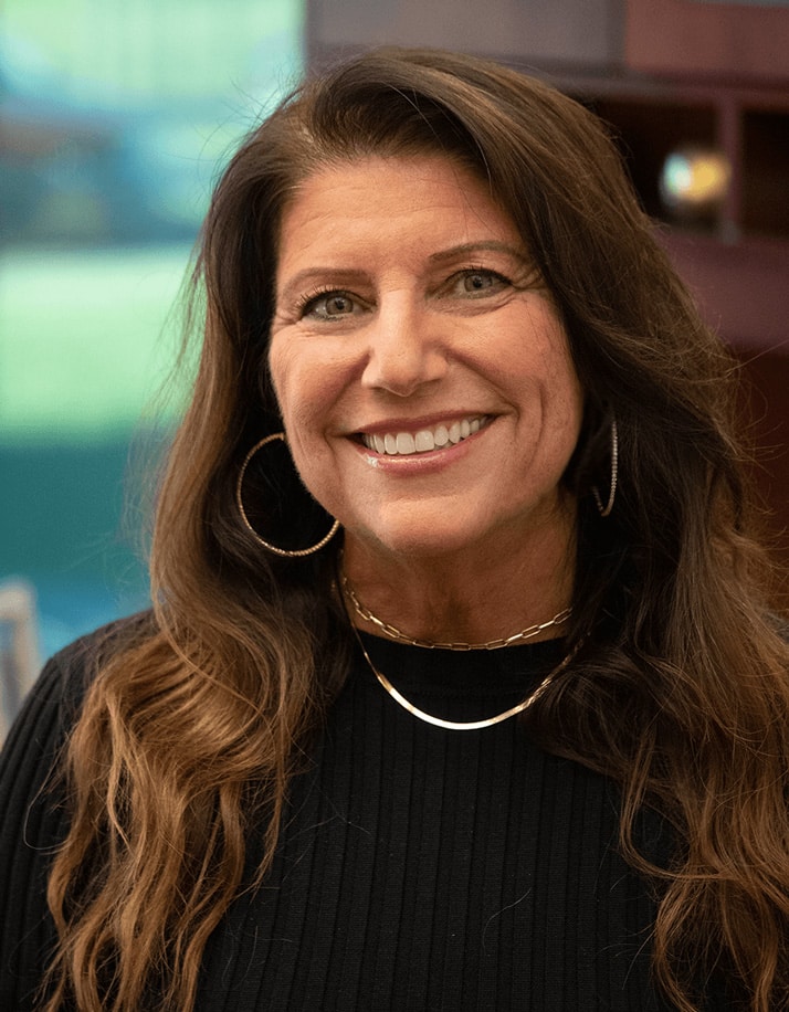 Woman with long brown hair and hoop earrings smiling while wearing a black top and gold jewelry.