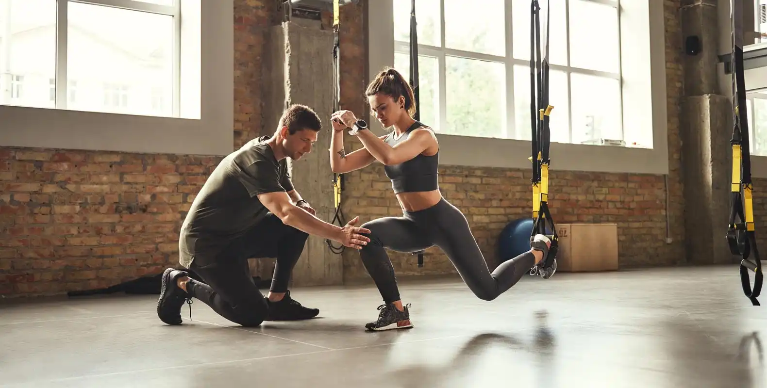 A woman exercises with suspension straps while a man kneels beside her, offering guidance in a gym.