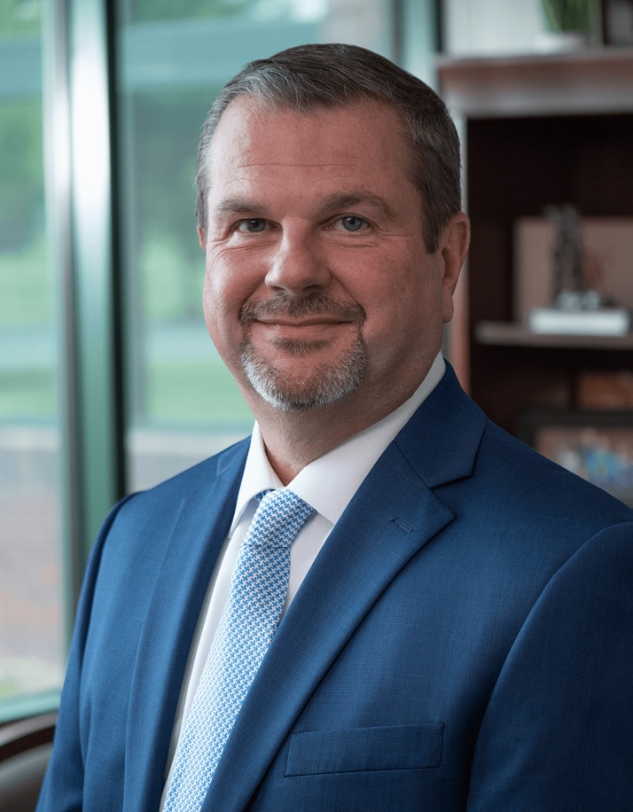 A man with short hair, a goatee, and a blue suit smiles while standing in front of an office window.