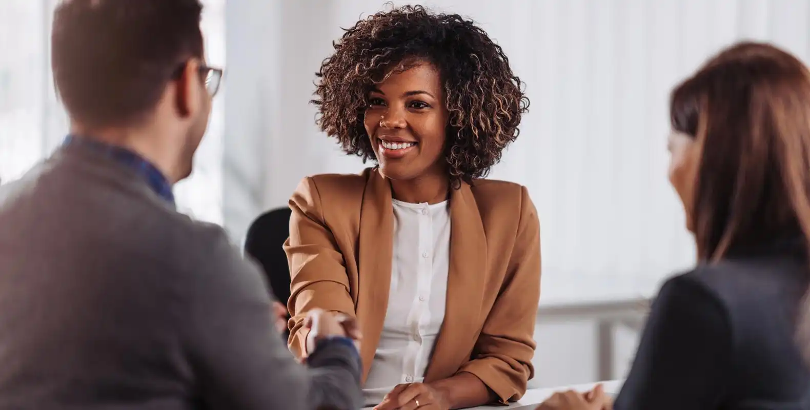 A woman in a beige blazer smiles and shakes hands with a man while another woman sits beside them.