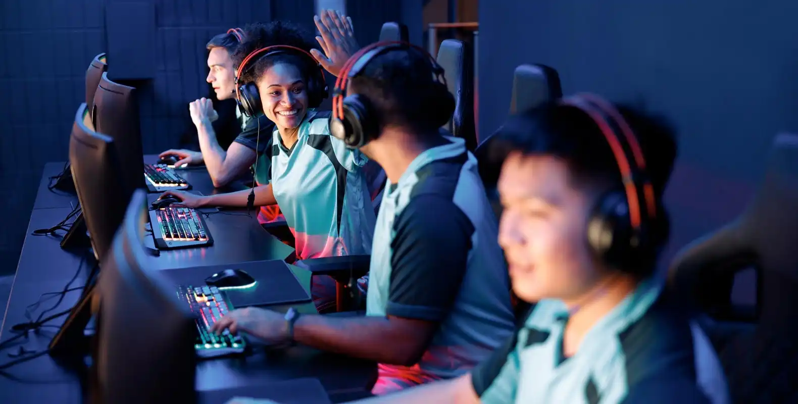 Four gamers wearing headsets are sitting and interacting at a row of computer stations, with one person high-fiving.