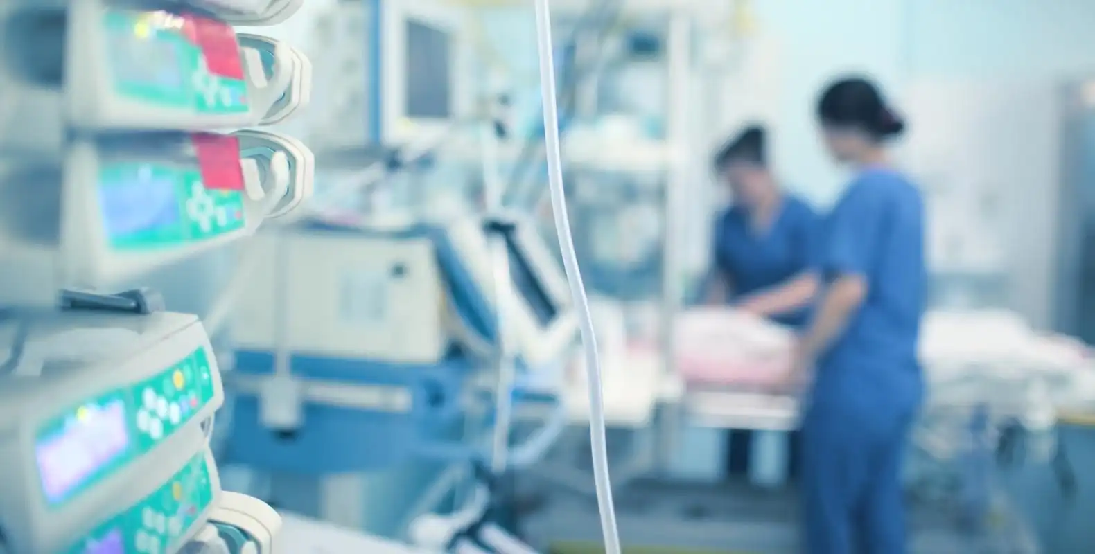 Hospital room with medical equipment in the foreground; two healthcare professionals work with a patient bed in the background.